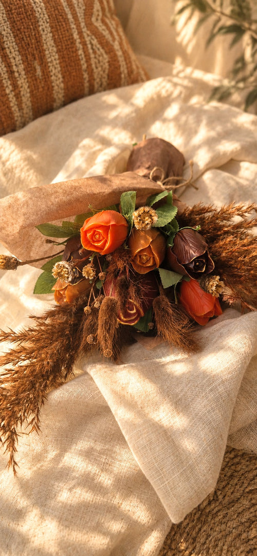 Bouquet Ambre Sauvage posé sur un drap écru : roses orangées et cuivrées, rose brun profond et pampa beige