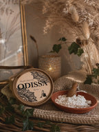 Boîte ronde Odissa Poudre Parfumée Dulcia posée sur toile de jute, avec coupelle en terre cuite remplie de poudre blanche, petite pelle en bois, bougeoir doré ajouré allumé et herbes séchées en arrière-plan
