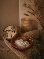 Boîte ouverte Odissa Poudre Parfumée Dulcia sur tissu naturel, coupelle en terre cuite avec poudre blanche et pelle en bois, bougeoir doré ajouré et feuilles de lierre vert en décor chaud et tamisé