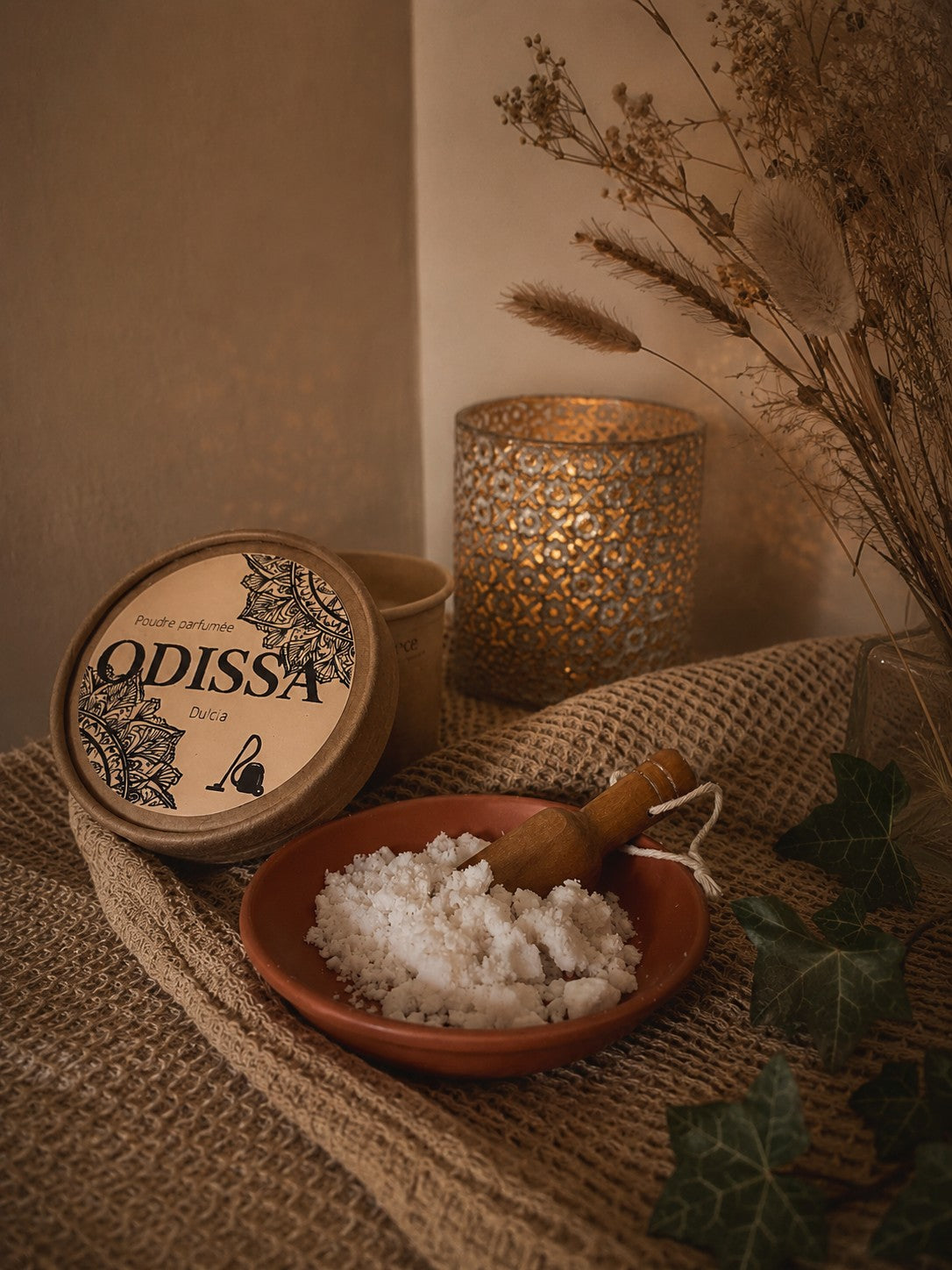 Boîte ouverte Odissa Poudre Parfumée Dulcia sur tissu naturel, coupelle en terre cuite avec poudre blanche et pelle en bois, bougeoir doré ajouré et feuilles de lierre vert en décor chaud et tamisé