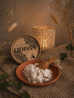 Poudre parfumée artisanale Odissa Dulcia dans une coupelle en terre cuite avec pelle en bois, boîte ronde à motifs mandala, bougeoir doré et lierre vert sur fond de graminées séchées et miroir doré
