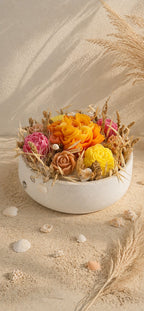 Composition florale Rivage d'Été en cire végétale, vue de côté de la vasque ronde couleur sable ponctuée de coquillages, avec les fleurs de cire orange, fuchsia et jaune