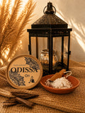 Poudre aspirateur parfumée Odissa Ori 100 g, boîte ronde avec poudre blanche dans une coupelle terracotta et cuillère en bois devant une lanterne orientale