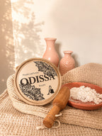 Boîte ronde Odissa Poudre Parfumée Poudré posée sur un tissu en coton naturel, avec un pilon en bois et une coupelle en terre cuite remplie de poudre blanche, vases roses en arrière-plan
