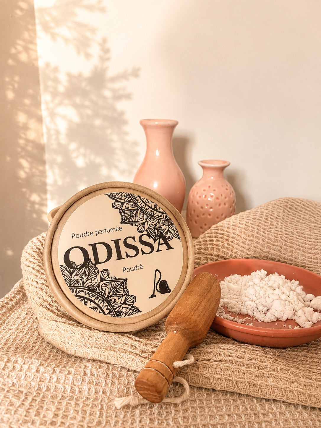 Boîte ronde Odissa Poudre Parfumée Poudré posée sur un tissu en coton naturel, avec un pilon en bois et une coupelle en terre cuite remplie de poudre blanche, vases roses en arrière-plan