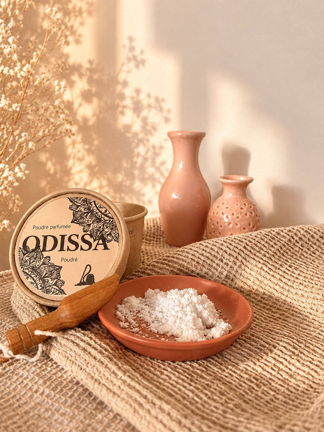 Boîte ouverte de poudre parfumée Odissa Poudré avec pilon en bois, coupelle en terre cuite et poudre blanche, décor naturel avec fleurs séchées et vases roses