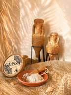 Poudre parfumée artisanale Odissa Azur présentée dans une coupelle en terre cuite avec pelle en bois, boîte ronde à motifs mandala, deux amphores texturées sur support métal et feuille de palmier séchée en arrière-plan