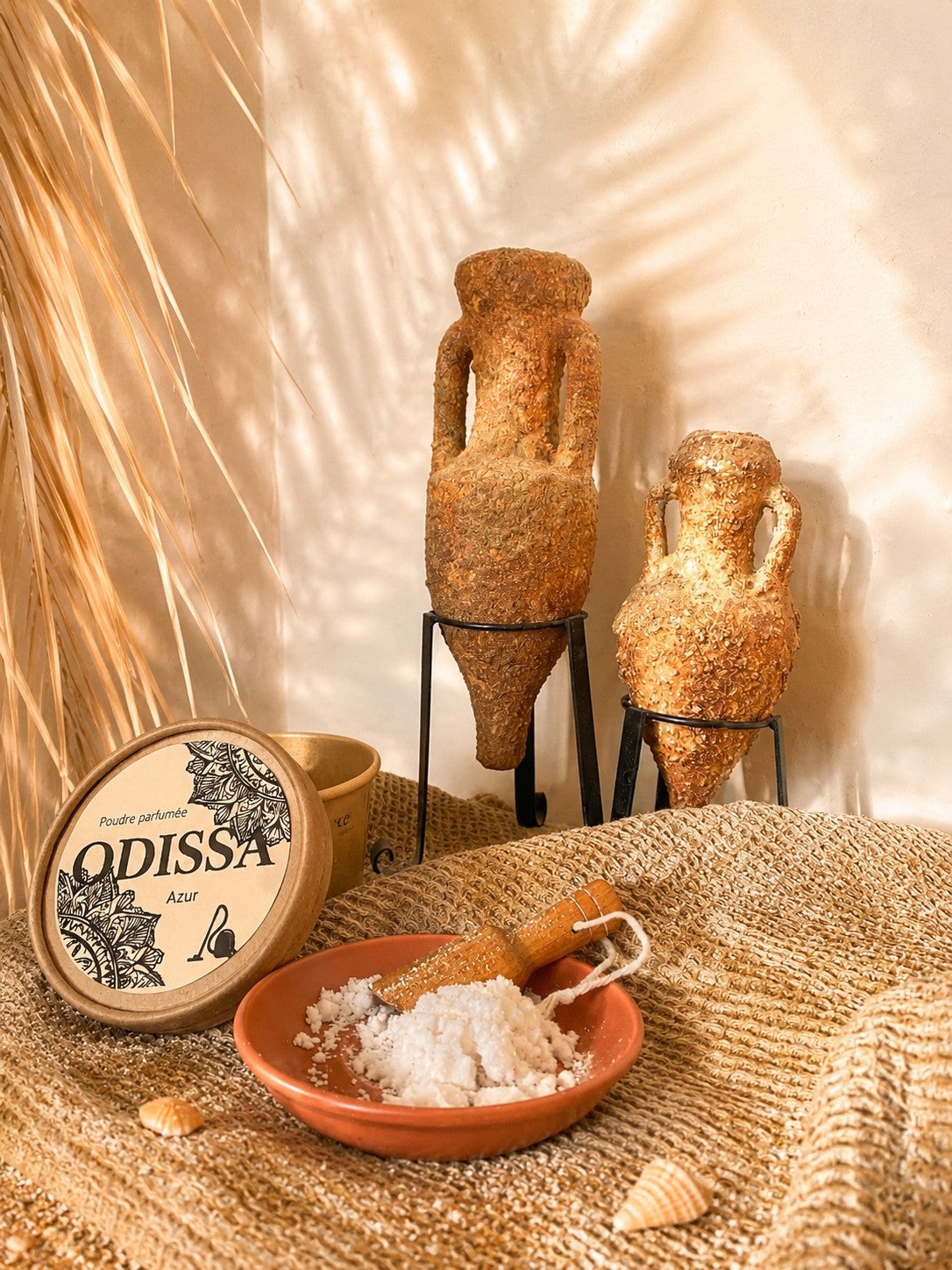 Poudre parfumée artisanale Odissa Azur présentée dans une coupelle en terre cuite avec pelle en bois, boîte ronde à motifs mandala, deux amphores texturées sur support métal et feuille de palmier séchée en arrière-plan