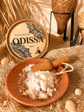Boîte ronde Odissa Poudre Parfumée Azur posée sur toile de jute avec coupelle en terre cuite remplie de poudre blanche, petite pelle en bois, coquillages et amphores texturées sur fond doré chaud