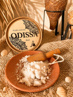 Boîte ronde Odissa Poudre Parfumée Azur posée sur toile de jute avec coupelle en terre cuite remplie de poudre blanche, petite pelle en bois, coquillages et amphores texturées sur fond doré chaud