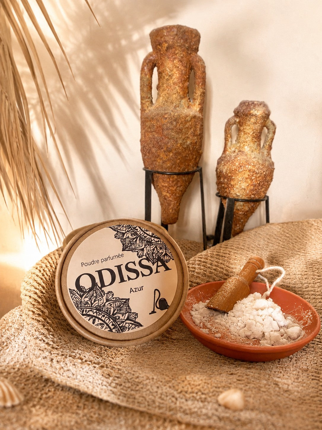 Boîte Odissa Poudre Parfumée Azur fermée sur tissu naturel, coupelle en terre cuite avec poudre blanche et pelle en bois, amphores artisanales et ombre de feuille de palmier sur fond beige lumineux