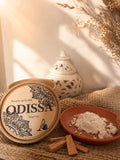 Boîte ouverte Odissa Poudre Parfumée Essence sur tissu naturel, avec coupelle en terre cuite, pilon en bois et bâtons de bois précieux, brûleur ajouré blanc et fleurs séchées en arrière-plan
