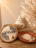 Poudre parfumée artisanale Odissa Essence présentée dans une coupelle en terre cuite, boîte ronde ouverte avec bâtons de bois précieux au premier plan, brûleur blanc et herbes séchées en décor