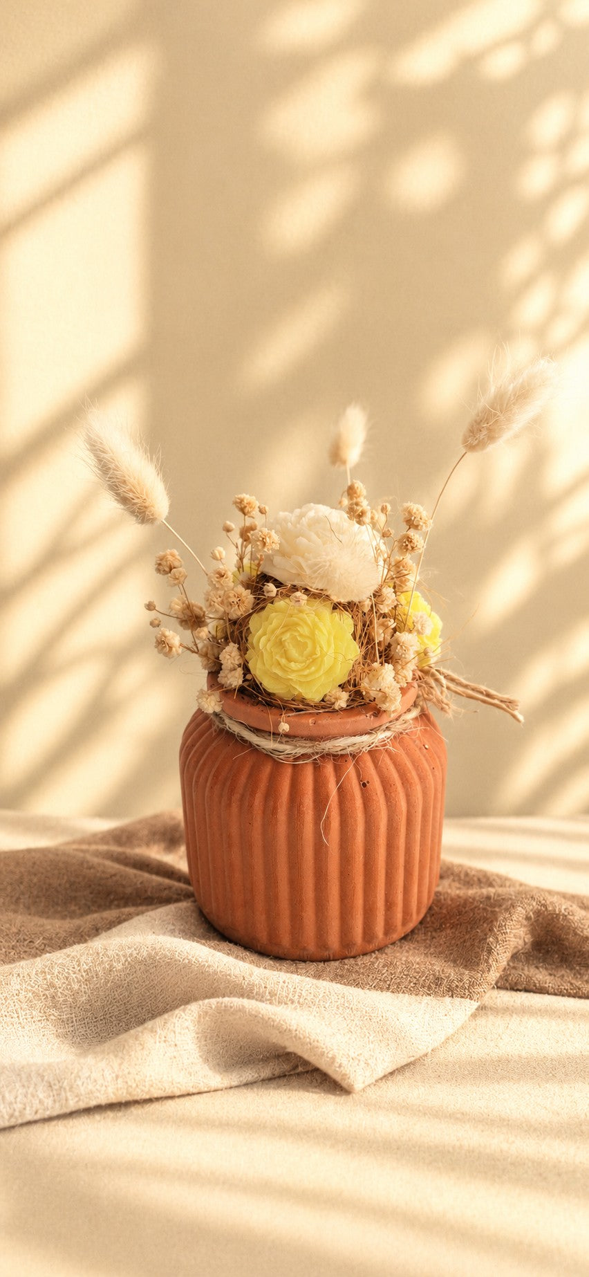 Composition florale en cire végétale avec roses jaune et crème, fleurs séchées et plumeaux dans un vase terracotta strié